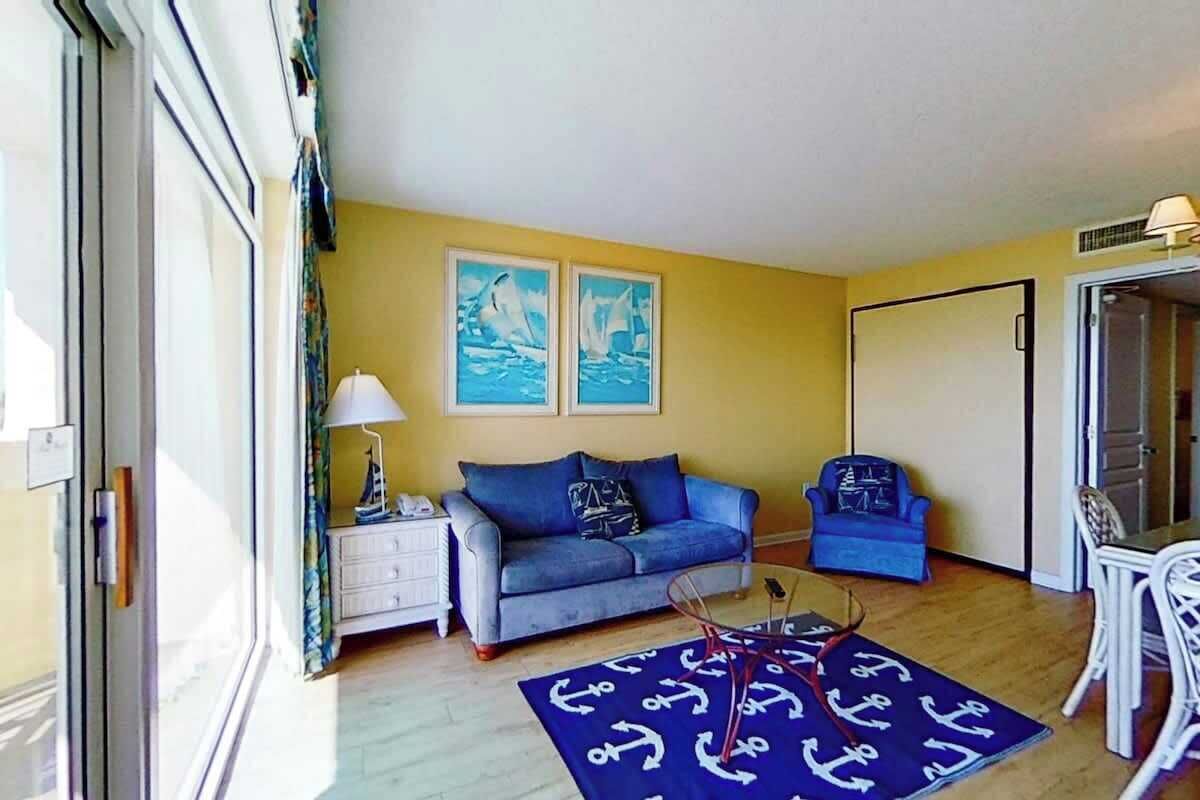 Habitación con sofá, una silla, una mesita y alfombra con motivos de anclas. Hay dos cuadros en la pared y una lámpara sobre una mesita auxiliar.