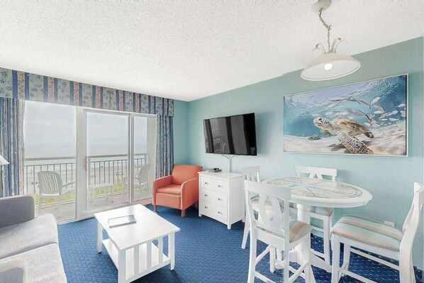 Ein Zimmer mit Balkon, Fernseher, Esstisch mit Stühlen und einem Gemälde einer Meeresschildkröte.