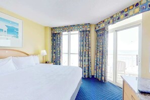 A hotel room with a large bed, a desk, a chair, and a balcony with curtains. - Bay Watch Resort 0505 (North Myrtle Beach)