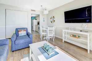 A living room with a TV, a sofa, a dining table, and a kitchen in the background. - Bay Watch Resort 0436 Oceanfront (North Myrtle Beach)