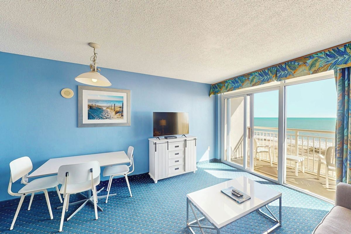 Habitación con pared azul, una mesa blanca, televisor y balcón con vista a la playa.