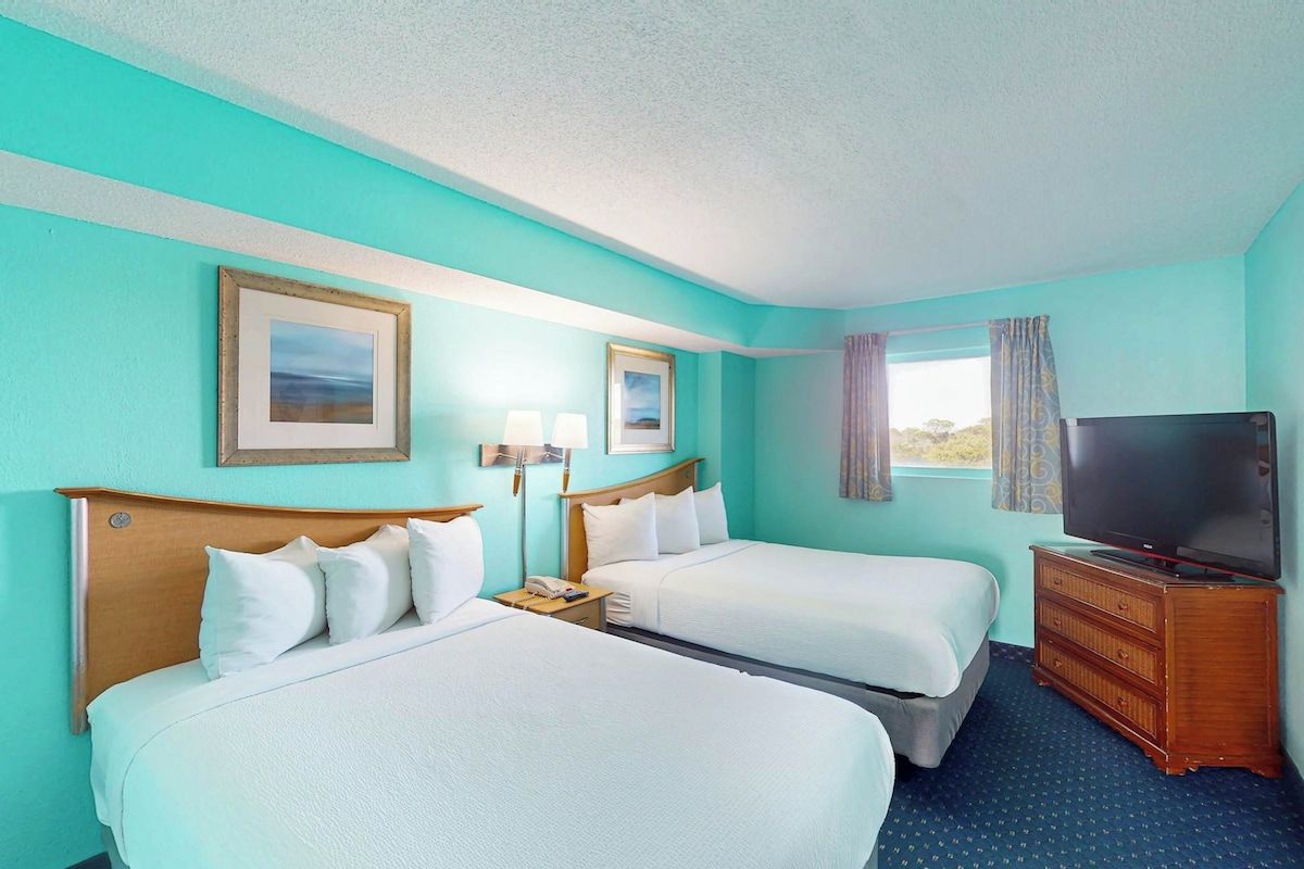 A hotel room with two beds, a television, a dresser, and two wall-mounted lamps.
