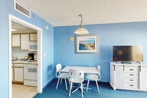 A room with a blue wall, a white table with four chairs, a television, and a framed picture on the wall. - Bay Watch Resort 0507 (North Myrtle Beach)