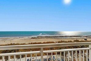 Blick auf den Strand vom Balkon aus, mit Geländer, blauem Himmel und Meer im Hintergrund.
