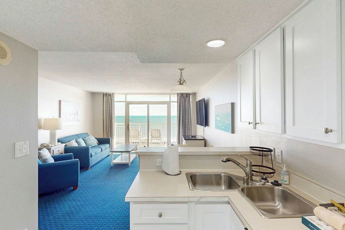 A compact hotel room with a kitchenette, a seating area with a blue sofa, a large window with a view, and a ceiling fan.