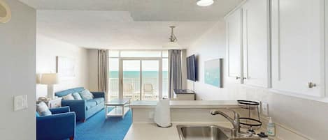 A compact hotel room with a kitchenette, a seating area with a blue sofa, a large window with a view, and a ceiling fan.