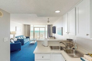 A compact hotel room with a kitchenette, a seating area with a blue sofa, a large window with a view, and a ceiling fan. - Bay Watch Resort 0513 (North Myrtle Beach)