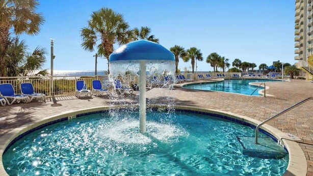 A pool area with a water feature, lounge chairs, and a building in the background. - Bay Watch Resort 0513 (North Myrtle Beach)