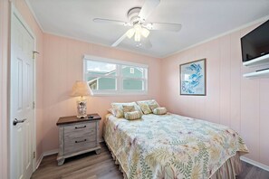 A bedroom with a bed, a nightstand, a lamp, a ceiling fan, a window, and a TV. - Blue Marlin (Galveston)