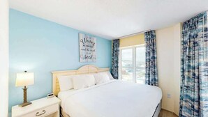 Una habitación de hotel con cama, mesita de noche con lámpara, pared con una cita motivacional y ventana con cortinas.