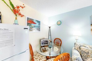 A compact living space with a glass dining table, wooden chairs, a refrigerator, a clock, and a lamp. - Bay Watch Resort 0531 (North Myrtle Beach)