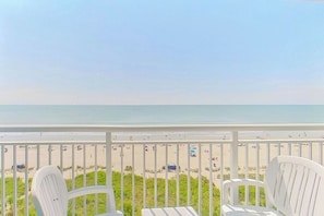 A balcony with white chairs overlooking a beach with people and umbrellas. - Bay Watch Resort 0620 (North Myrtle Beach)
