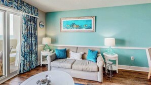 Ein Zimmer mit einer türkisfarbenen Wand, einem beigen Sofa mit blauen und weißen Kissen, einem weißen Couchtisch und einem gerahmten Gemälde über dem Sofa.