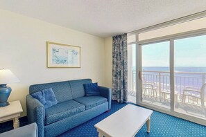 Ein Zimmer mit einem blau gemusterten Sofa, einem weißen Tisch und einem großen Fenster mit Meerblick.