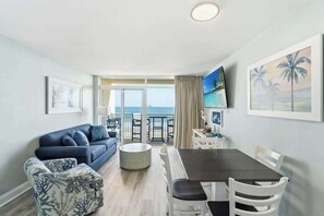 A room with a sofa, dining table, chairs, a TV, and a balcony with a view of the sea. - Bay Watch Resort 0603 (North Myrtle Beach)