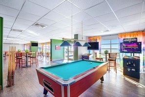 A pool table in a brightly lit indoor space with a green pool table, a pool cue rack, and a billiards table in the background. - Bay Watch Resort 0603 (North Myrtle Beach)