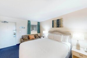 A hotel room with a large bed, a bedside table with a lamp, a small sofa, and a window with curtains. - Bay Watch Resort 0534 (North Myrtle Beach)