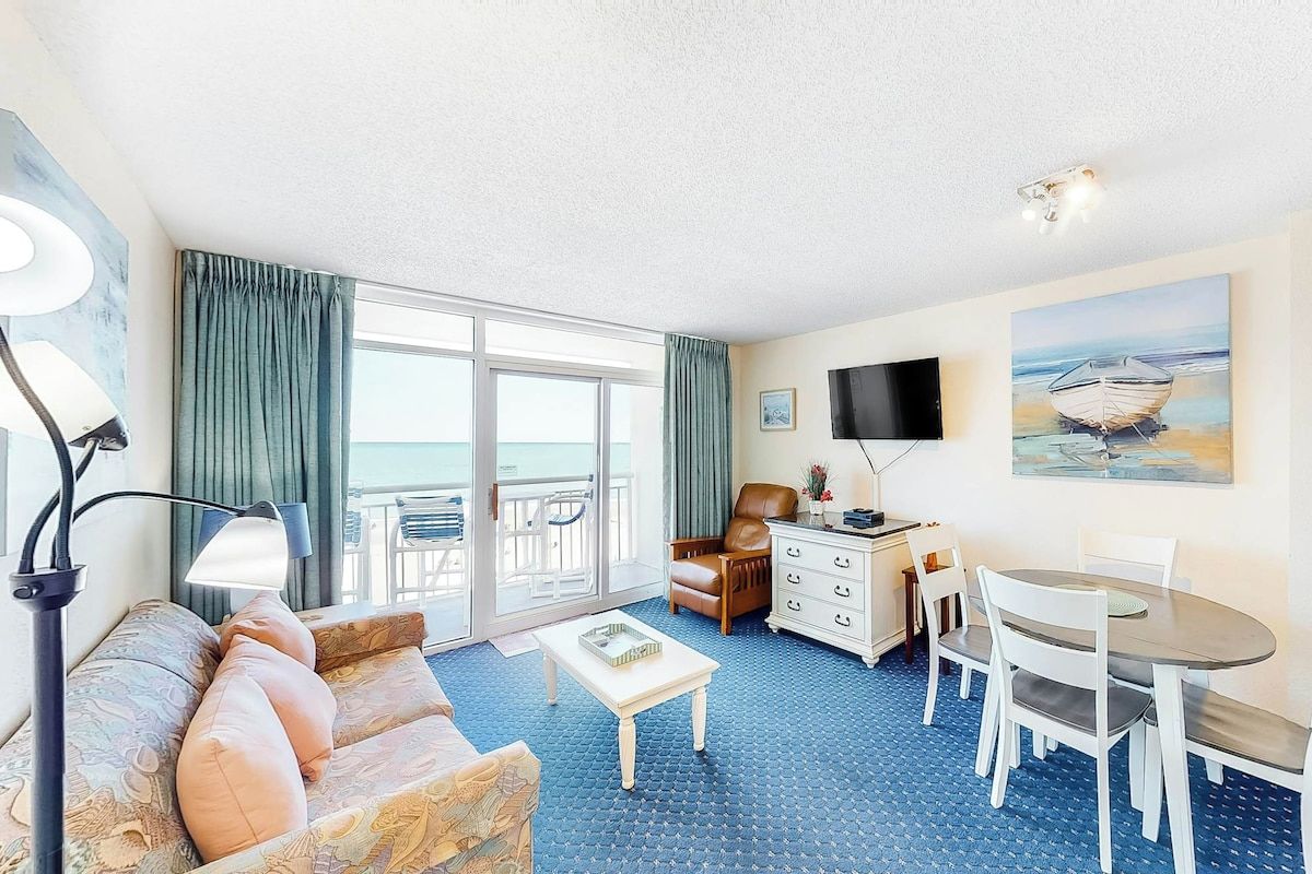 A hotel room with a sofa, a dining table, a TV, and a view of the sea through the window.