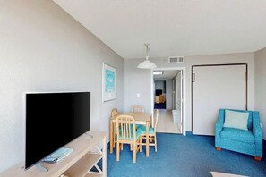 A room with a flat-screen TV, a dining table with chairs, a blue carpet, and a blue sofa. - Bay Watch Resort 0606 (North Myrtle Beach)