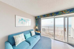 A room with a blue sofa, a framed artwork, a balcony with a view of the sea, and a tropical-themed curtain. - Bay Watch Resort 0606 (North Myrtle Beach)