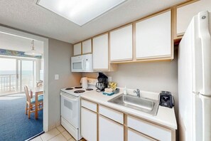 A compact kitchen with a microwave, stove, sink, and refrigerator. There is a dining area with a table and chairs visible through an open door. - Bay Watch Resort 0606 (North Myrtle Beach)