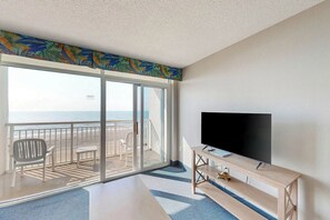A room with a large sliding glass door leading to a balcony with a view of the ocean. There is a flat-screen TV on a wooden stand and a small table with a chair. - Bay Watch Resort 0606 (North Myrtle Beach)