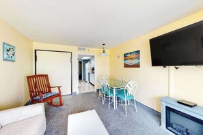 A living room with a sofa, a wooden chair, a dining table with chairs, a flat-screen TV, and a fireplace. - Bay Watch Resort 0604 (North Myrtle Beach)