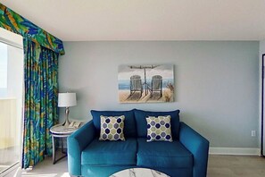 A room with a blue sofa, two patterned cushions, a painting of beach chairs, a lamp, and a window with curtains. - Bay Watch Resort 0607 (North Myrtle Beach)