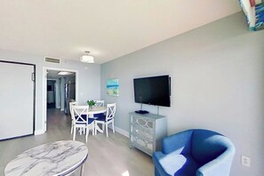 Ein Wohnzimmer mit Fernseher, einem blauen Sessel, einem Esstisch mit Stühlen und einem Marmor-Couchtisch.