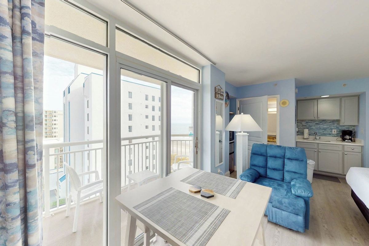 A hotel room with a balcony, a dining table, a sofa, and a kitchenette.