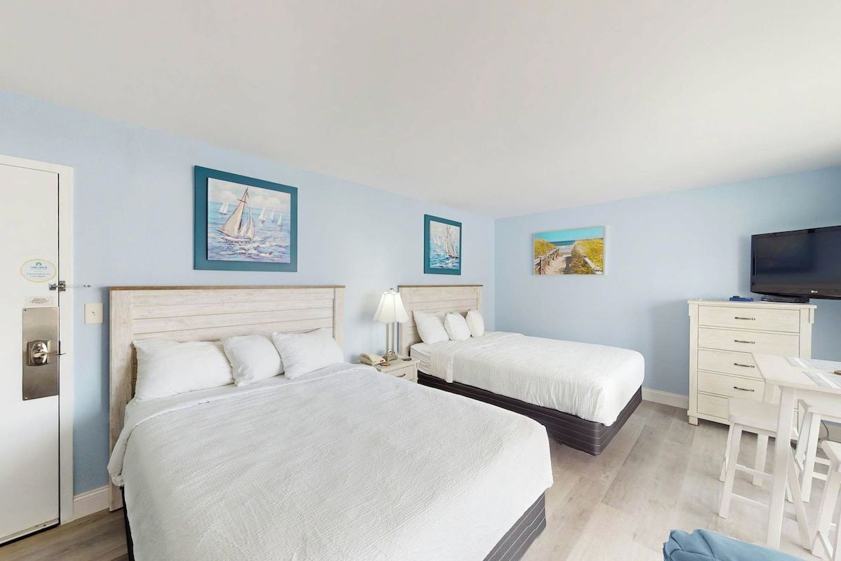 Habitación de hotel con dos camas, un escritorio, una televisión y obras de arte en las paredes.