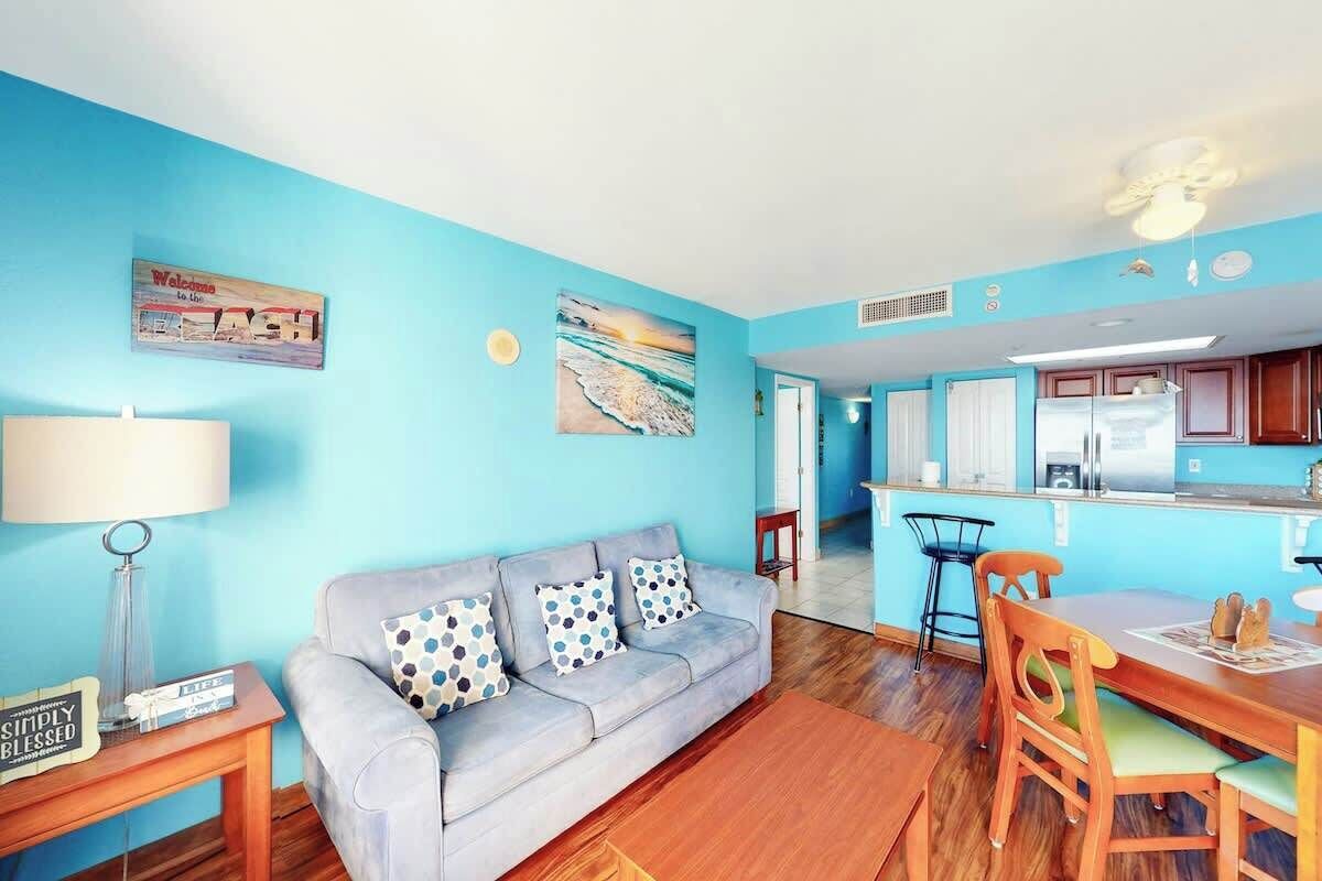 A living room with a sofa, a dining table with chairs, a kitchen area, and a ceiling fan.