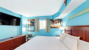 Ein Hotelzimmer mit einem großen Bett, einem an der Wand montierten Flachbildfernseher, einer Holzkommode und einem Bild mit einer Strandansicht.