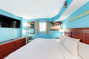 Ein Hotelzimmer mit einem großen Bett, einem an der Wand montierten Flachbildfernseher, einer Holzkommode und einem Bild mit einer Strandansicht.
