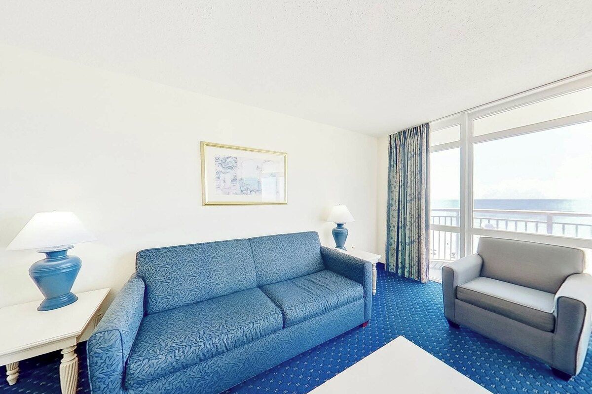 Habitación de hotel con un sofá azul estampado, un sillón gris, una mesita de noche con lámpara y un ventanal con cortinas.