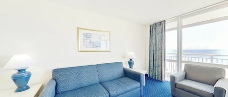 A hotel room with a blue patterned sofa, a grey armchair, a small table with a lamp, and a large window with curtains.