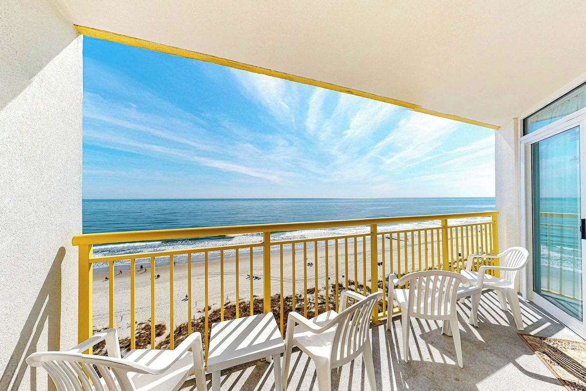 A balcony with a view of the beach and ocean, white plastic chairs, and a yellow railing.
