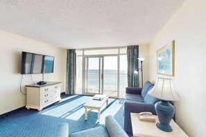 A hotel room with a television, a sofa, a table, a lamp, and a view of the sea through large windows. - Bay Watch Resort 0714 Oceanfront (North Myrtle Beach)