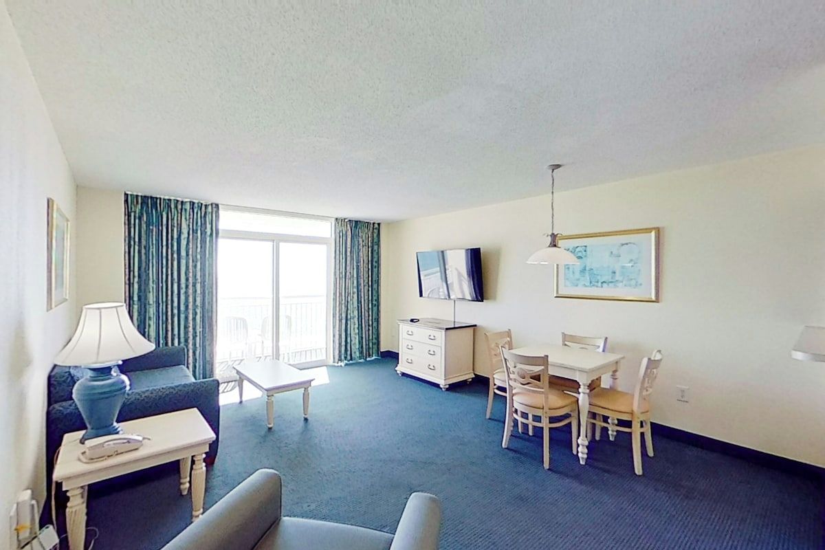 Ein Hotelzimmer mit einer Couch, einem kleinen Tisch mit Lampe, einem Fernseher, einem Esstisch mit Stühlen und einem Fenster mit Vorhängen.
