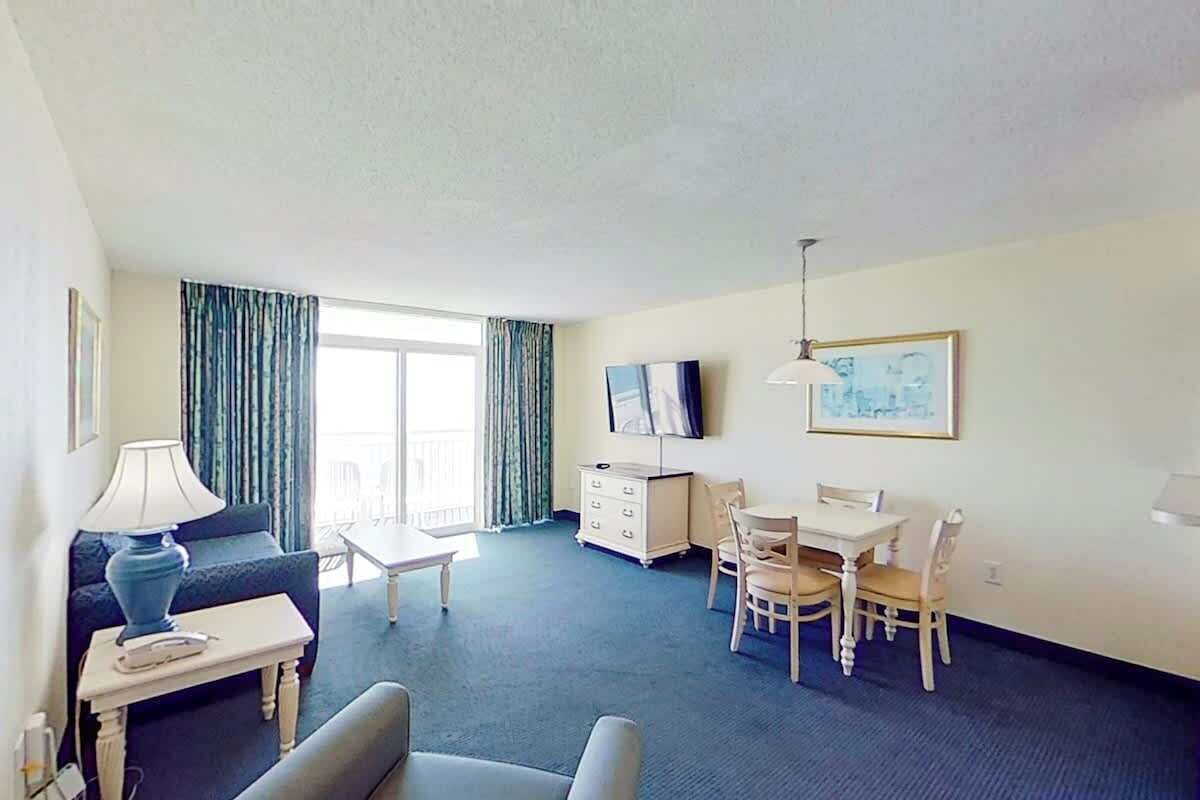 Ein Hotelzimmer mit einer Couch, einem kleinen Tisch, einem Fernseher, einem Esstisch mit Stühlen und einem Fenster mit Vorhängen.