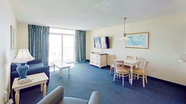 Ett hotellrum med en soffa, ett litet bord, en TV, ett matbord med stolar och ett fönster med gardiner.