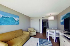 Uma sala de estar com sofá, poltrona, mesa de jantar com cadeiras, televisão e um quadro na parede.