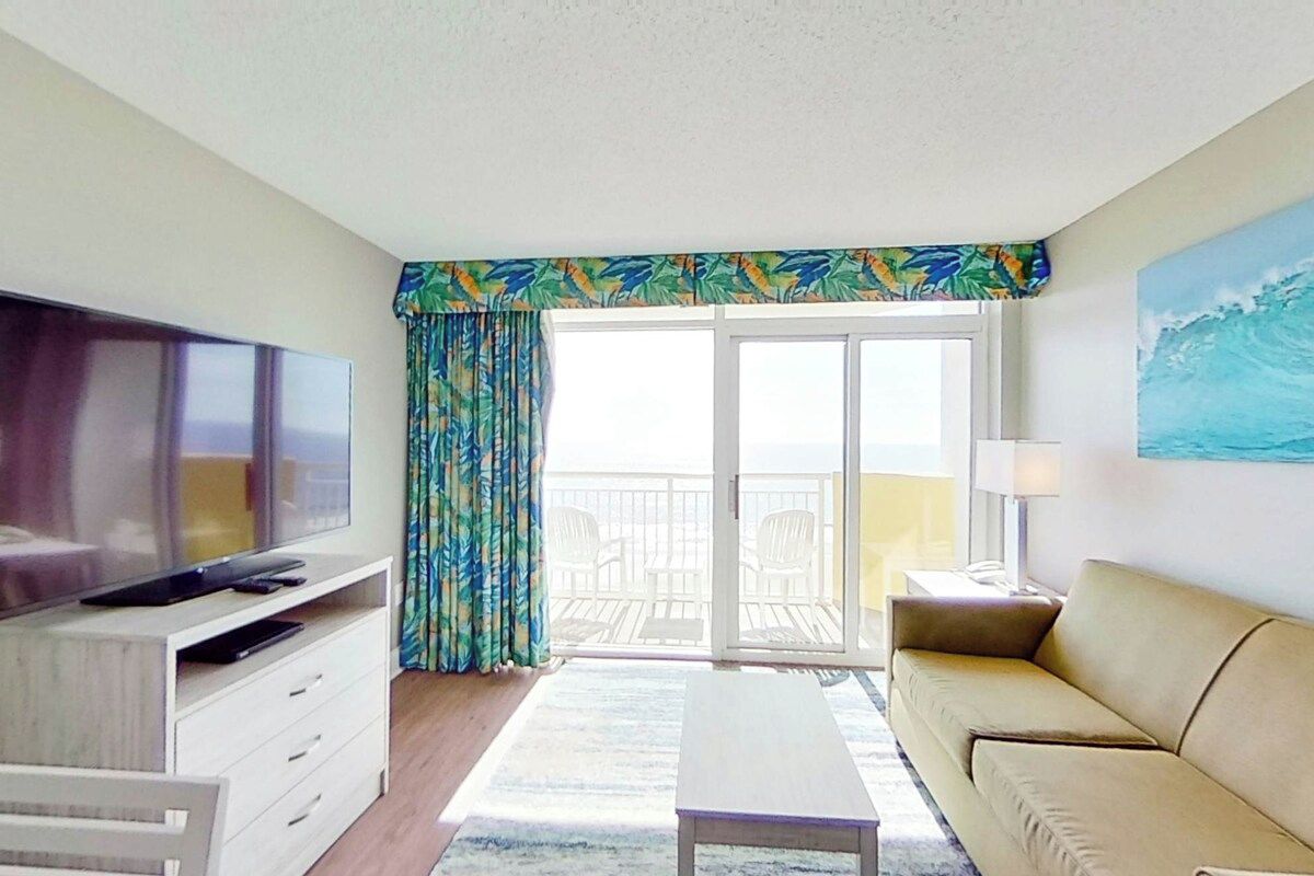 A hotel room with a television, a sofa, a coffee table, a balcony with a view, and curtains.