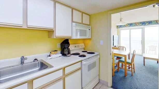 A compact kitchen with a microwave, stove, and sink, leading to a dining area with a table and chairs. - Bay Watch Resort 0807 (North Myrtle Beach)