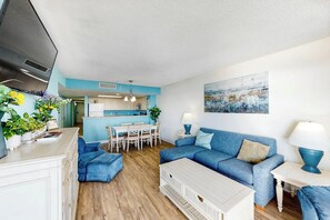 A modern living room with a blue sofa, a white coffee table, and a dining area with a table and chairs. - Bay Watch Resort 0815 (North Myrtle Beach)