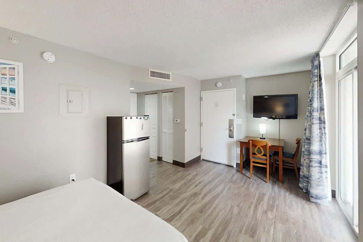 A hotel room with a bed, a kitchenette, a dining area with a table and chairs, a flat-screen TV, and a window with curtains.