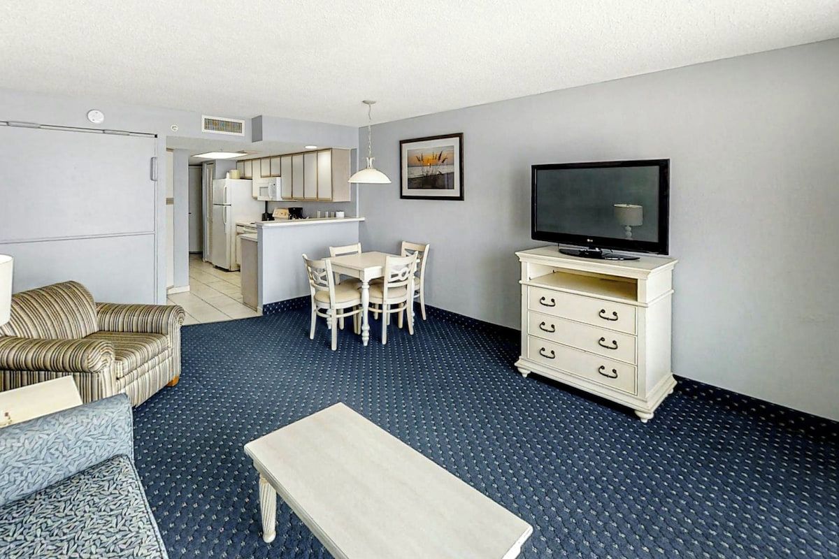 A compact living space with a dining area, kitchenette, sofa, TV, and a patterned floor.