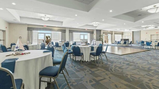 A spacious hotel conference room with round tables and blue chairs, large windows, and a carpeted floor. - Bay Watch Resort 0814 (North Myrtle Beach)