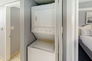 Una màquina de rentar compacta i blanca en una habitació amb un llit i un quadre a la paret.