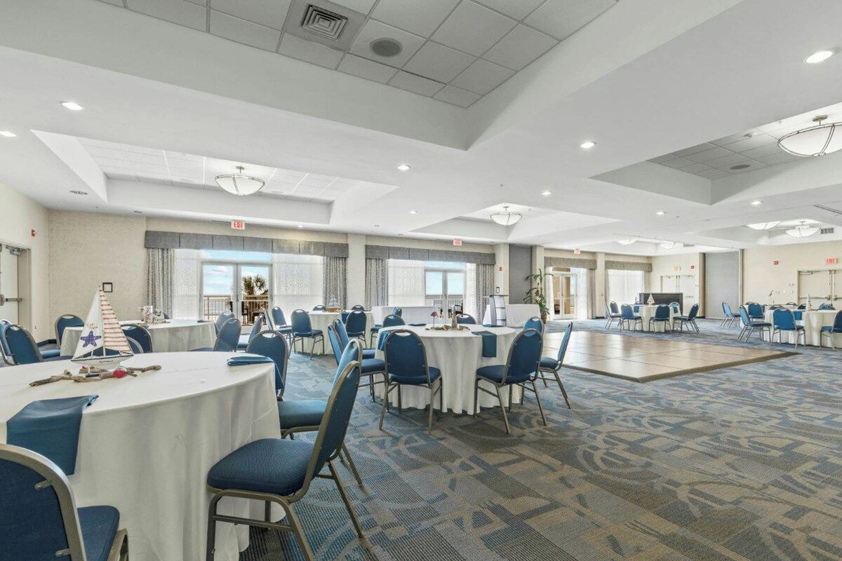 A hotel conference room with round tables, blue chairs, and a carpeted floor.